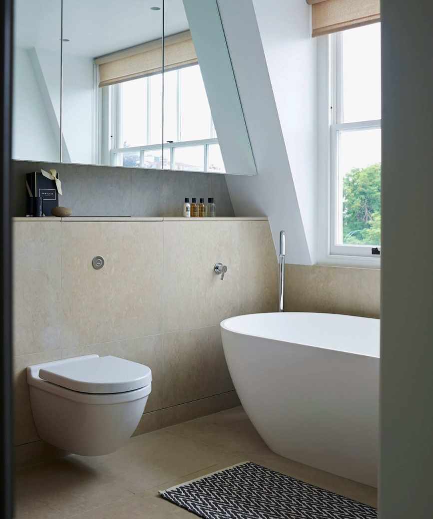 A floating toilet installed on a stone half-wall beside a free-standing bathtub. A large window above the bath lights the room.