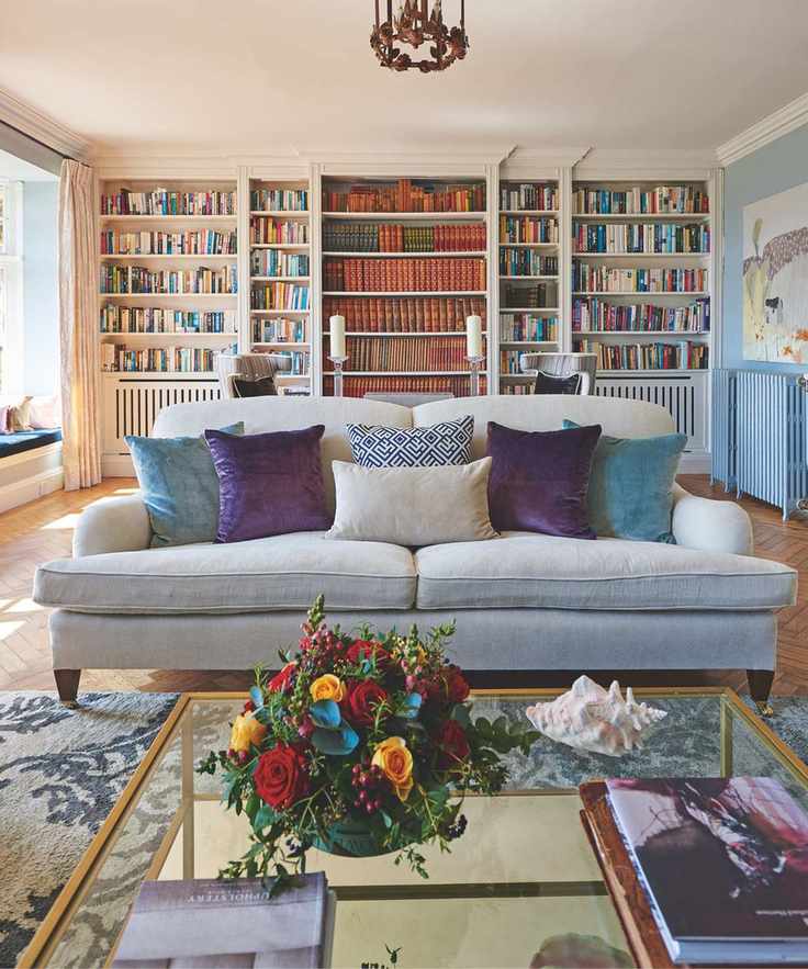 Colorful living room with white and blue walls, large white shelves filled with books, and a brass-look pendant lamp. In the foreground is a grey sofa with purple and blue velvet cushions, a large grey patterned rug, and a glass and gold coffee table with flowers, coffee table books and a white conch shell.