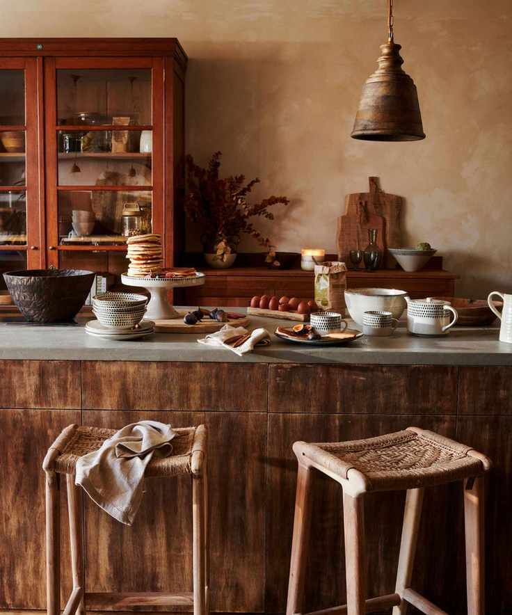 An earth toned kitchen, with wooden island and red painted pantry. Pancakes are stacked on a cake stand on the island. Two rattan stools pulled up.