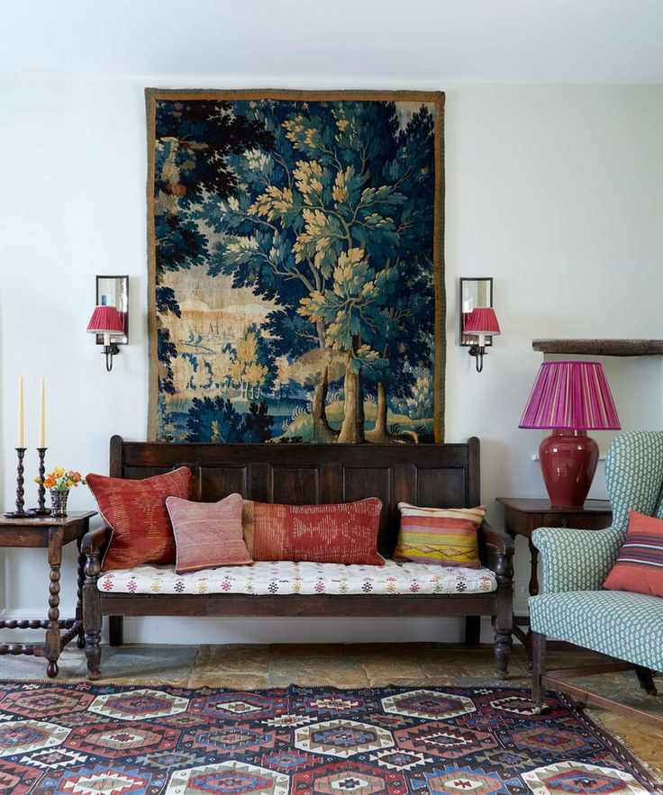 A rustic entryway featuring a dark wood settle bench with red pillows, a large tree-motif tapestry, and a vibrant geometric rug