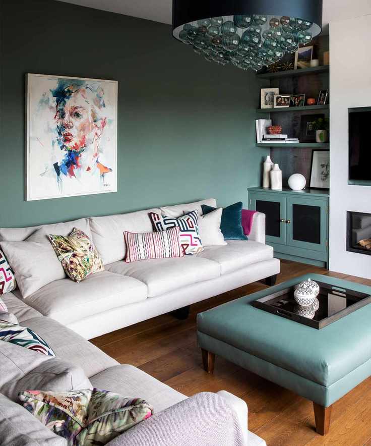 Living room with green walls and wooden floors featuring a large L-shaped gray sofa, with a green leather ottoman that matches the wall paint, and a striking pendant light with glass bulbs.