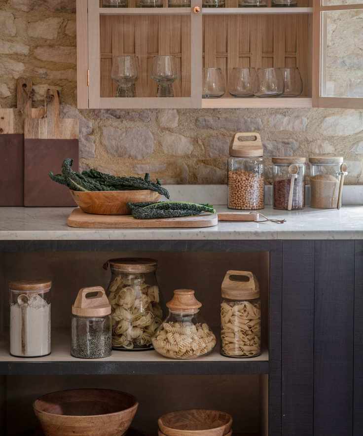 A rustic kitchen with white countertops, glass jars of dried food goods on the counter and shelves beneath, and dark blue painted cabinets.