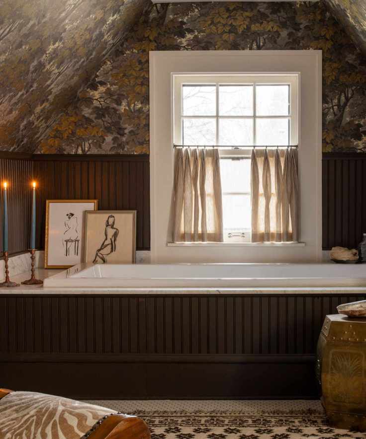 moody loft guest bathroom with dark wooden brown panelling and bath panel, small window with cafe curtain, botanical wallpaper, decorated with a rug, stool, artwork, candles and a small chair