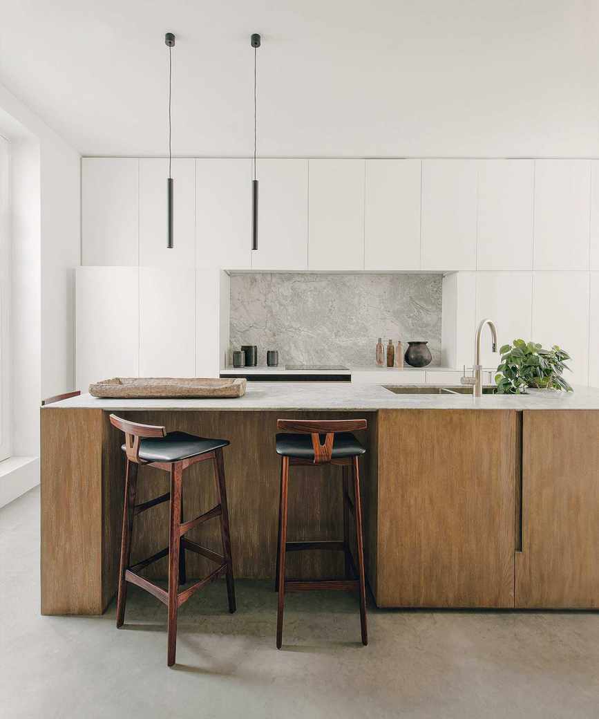 Modern kitchen space with kitchen island with dark wood base and cupboards, dark wood and black leather bar stools, white cupboards and white painted walls, two long black pendants hanging over island