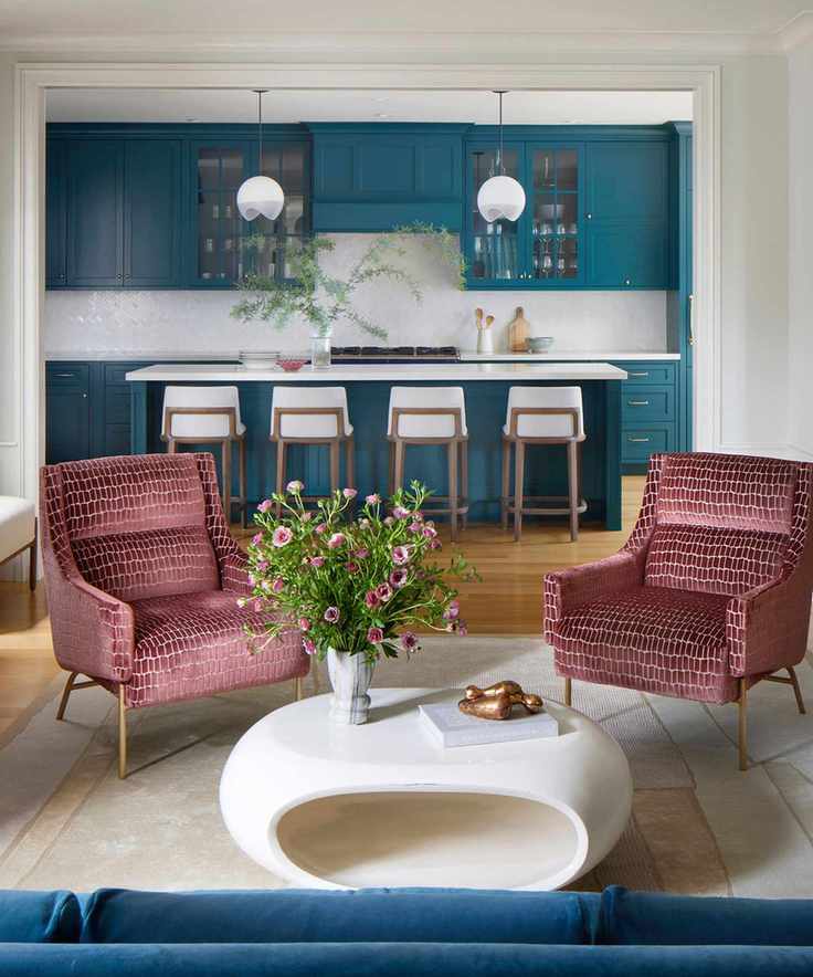 A cozy sitting area with red armchairs and a white coffee table, with a blue kitchen visible in the background.