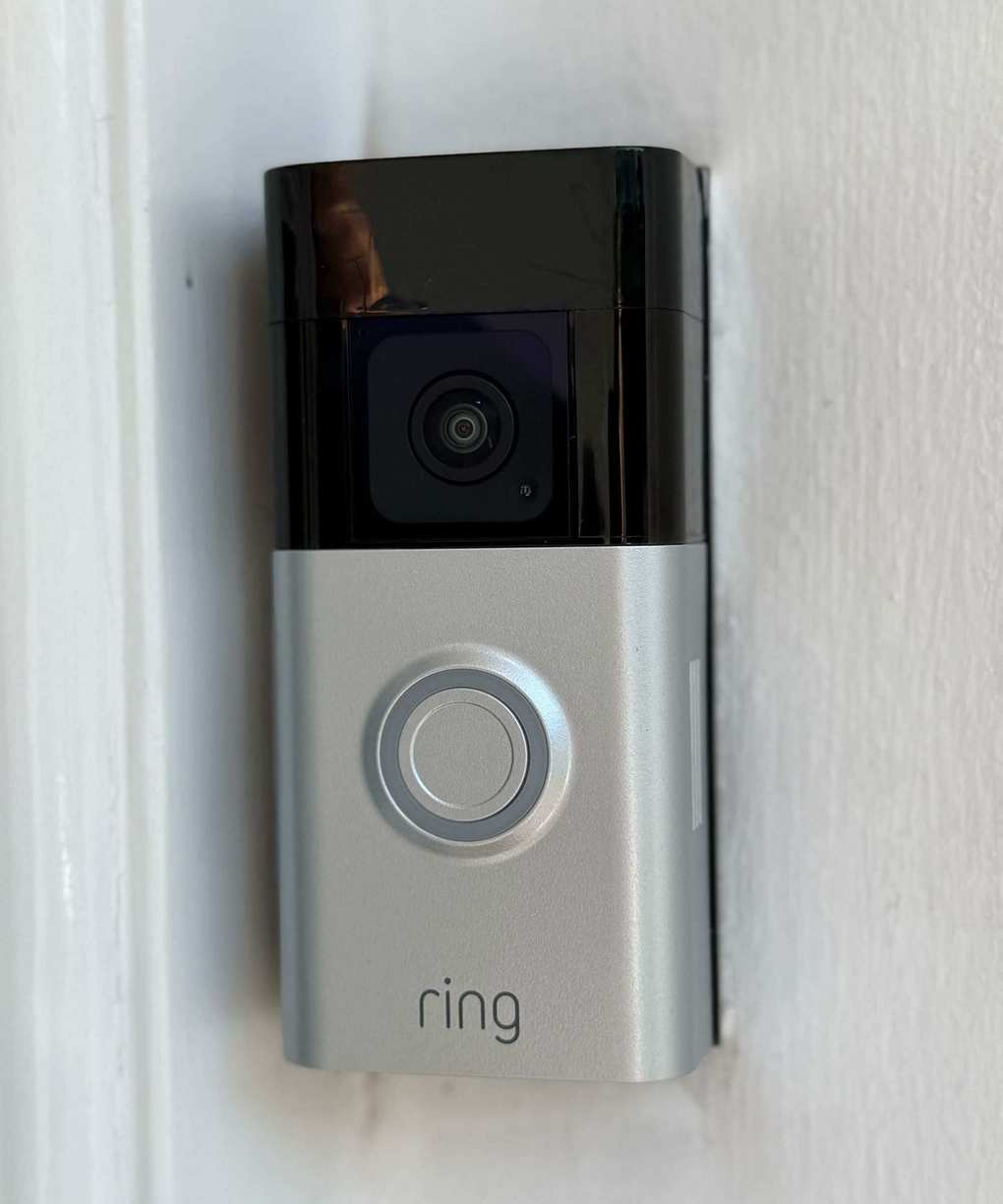 A black and silver Ring Battery Doorbell Plus mounted on a white painted wall. It features a camera lens, circular button, and Ring logo.
