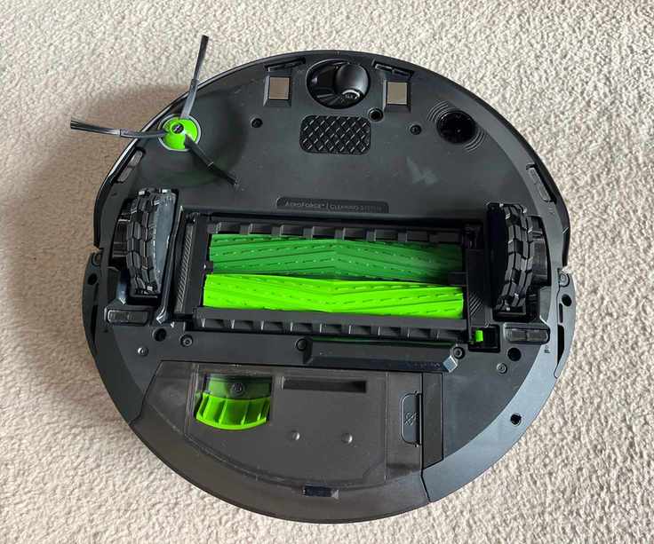 Underside of the iRobot Roomba Combo j9+ robot vacuum on cream carpet, showing bright green brushrolls, sweeping brush and black wheels