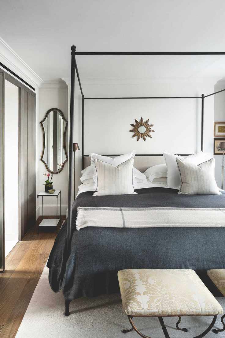 white bedroom with black four poster bed, black blanket, wooden floor, metal mirrors and side table, upholstered footstool and white rug