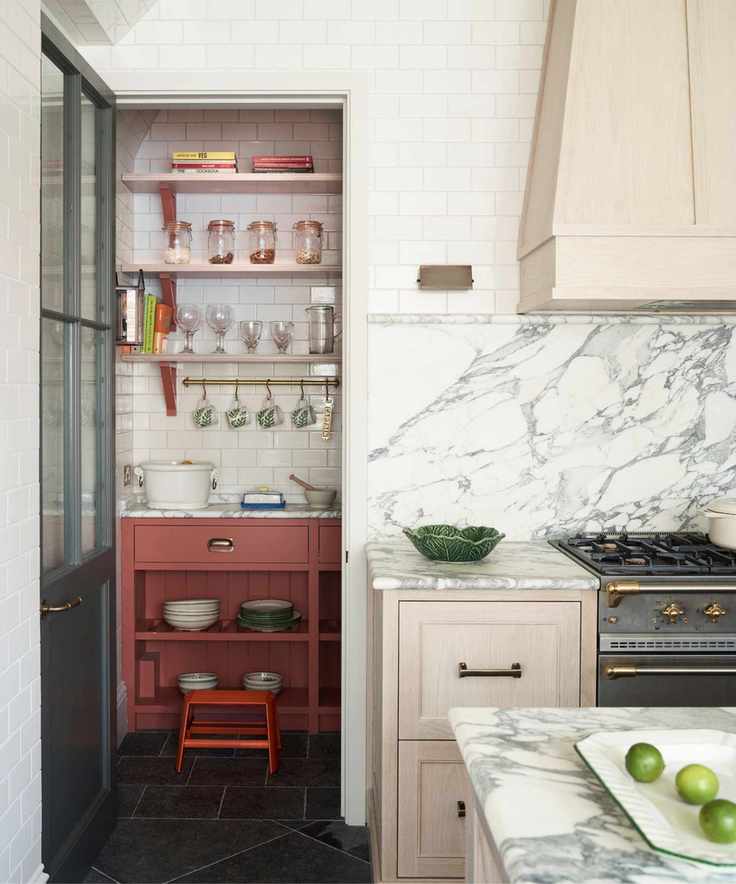 A marble kitchen with pink drawers and a range hood, featuring a grey hob and a white tiled pantry with pale red cabinets.