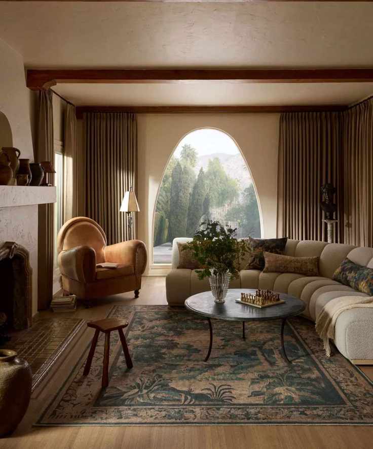 a living room with a large decorative rug, a curved gray couch, a tan accent chair, and an arched window framed by large tan drapes