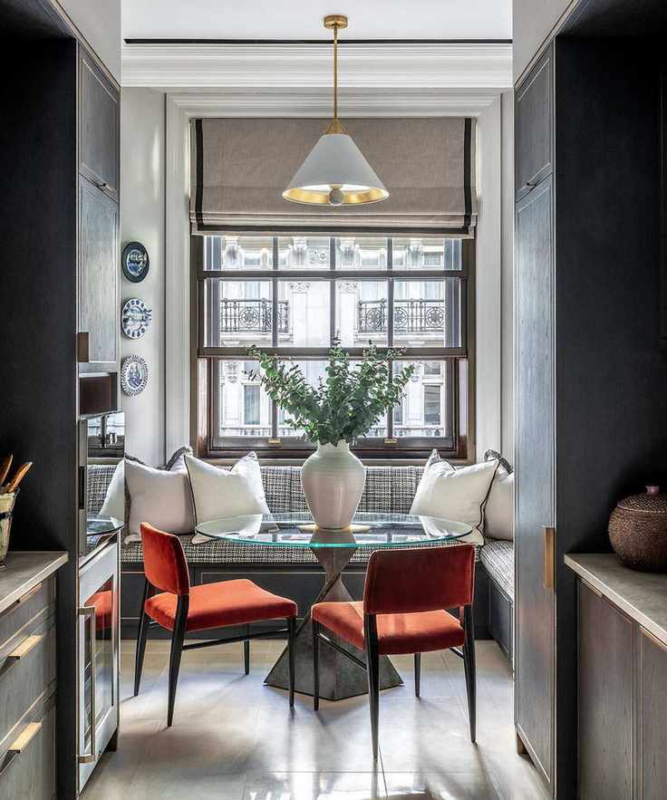 A round glass kitchen dining table in front of a banquette bench, with two red velvet chairs tucked beneath, illuminated by a large window at the end of a narrow kitchen.