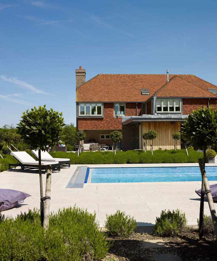 A pool area at the back of a house with a patio, sun loungers and flower beds with small trees