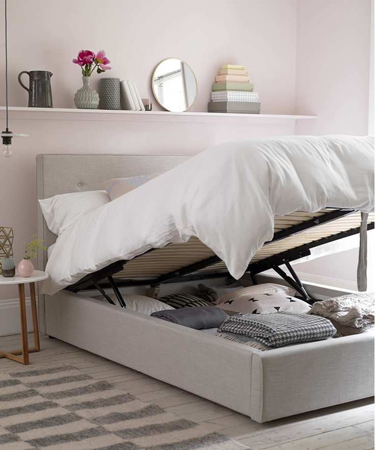 A grey fabric bed, lifted up with built-in underbed storage, filled with folded blankets and linens. White bedding on the duvet