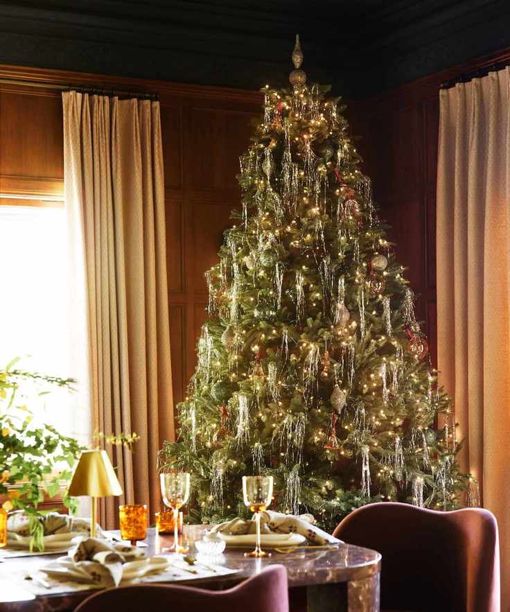 a wood-paneled dining room with a large Christmas tree covered in tinsel and various glass ornaments perched behind a dining table with glass tabletop details, plates, and utensils