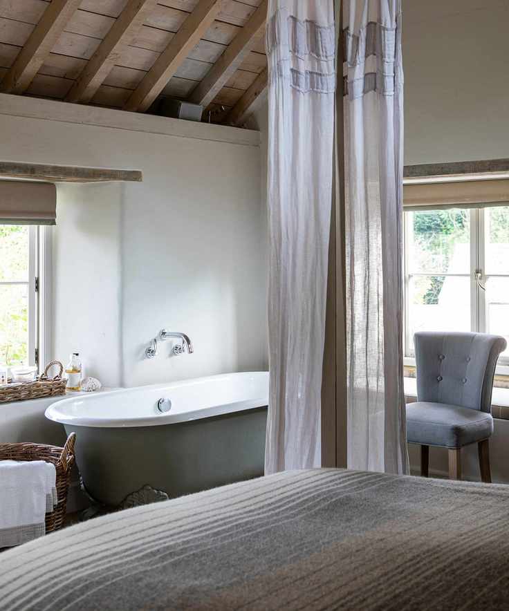 Traditional, country style bedroom with four poster bed with sheer curtains, small bath beside window, armchair beside other window, wooden vaulted ceiling