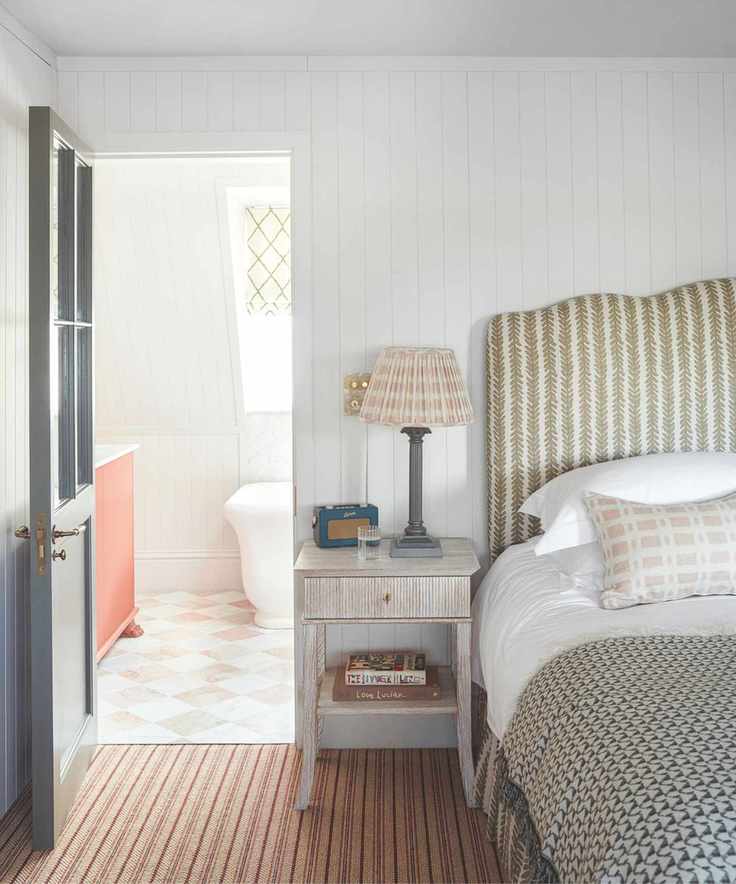 A white wood panelled bedroom with striped carpet floor, a double bed with green and white patterned headboard, wooden bedside table with lamp and radio, and a stack of books. To the left is a dark blue door, and behind a bright white and orange tiled bathroom with white bathtub is seen.