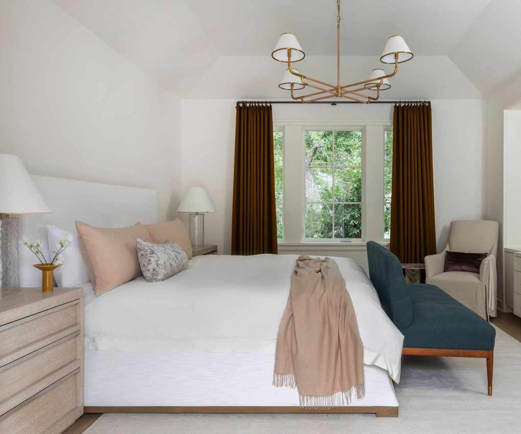 A bedroom with white walls, dark brown curtains, white bedding, a navy blue sofa at the end of the bed, a cream carpet, warm beige cushions, and a throw blanket.