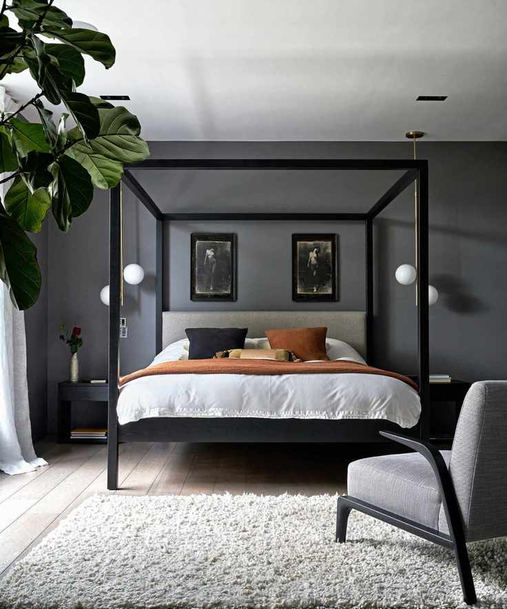 Gray bedroom with a white rug, a black four-poster bed with white bedding, cushions, a grey upholstered chair, and a large houseplant to the left.