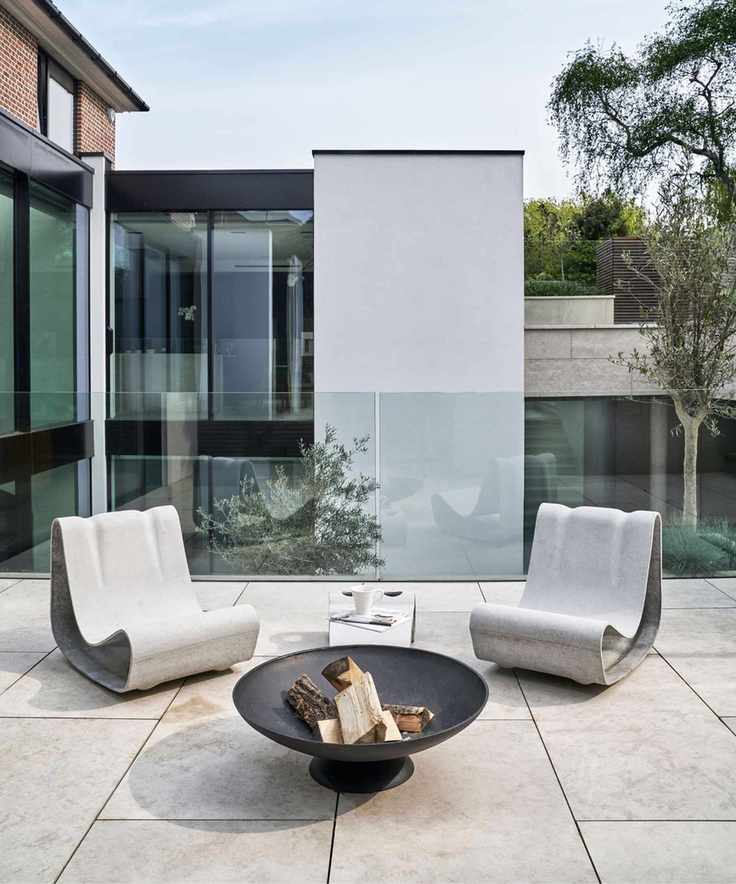 A concrete outdoor seating area with modern white chairs, a firepit filled with wood and a glass baluster
