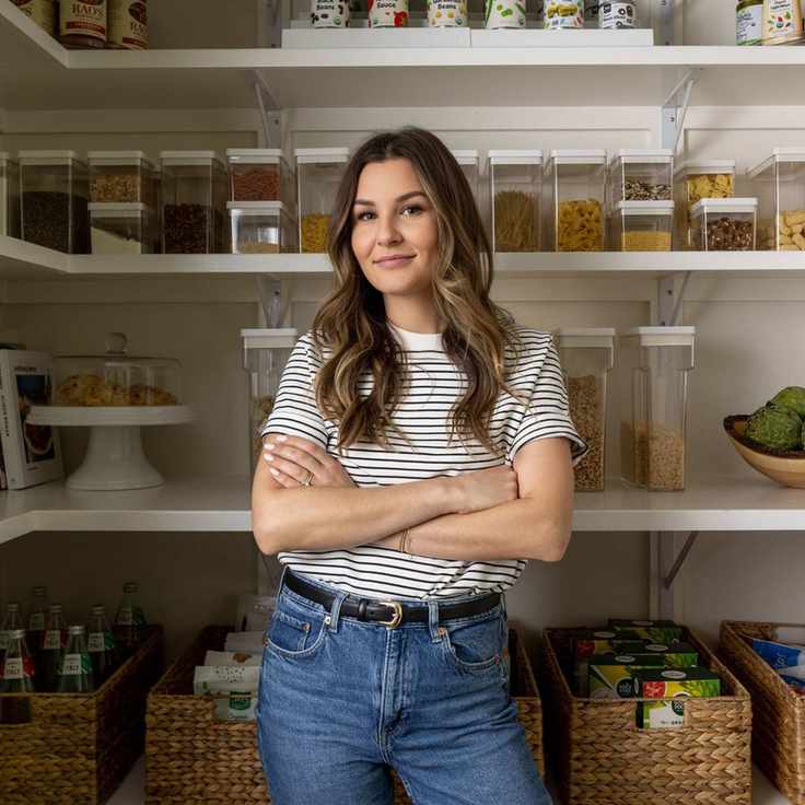 A picture of Ashley La Fond wearing a t-shirt and jeans, standing in a pantry with jars and cans behind her.