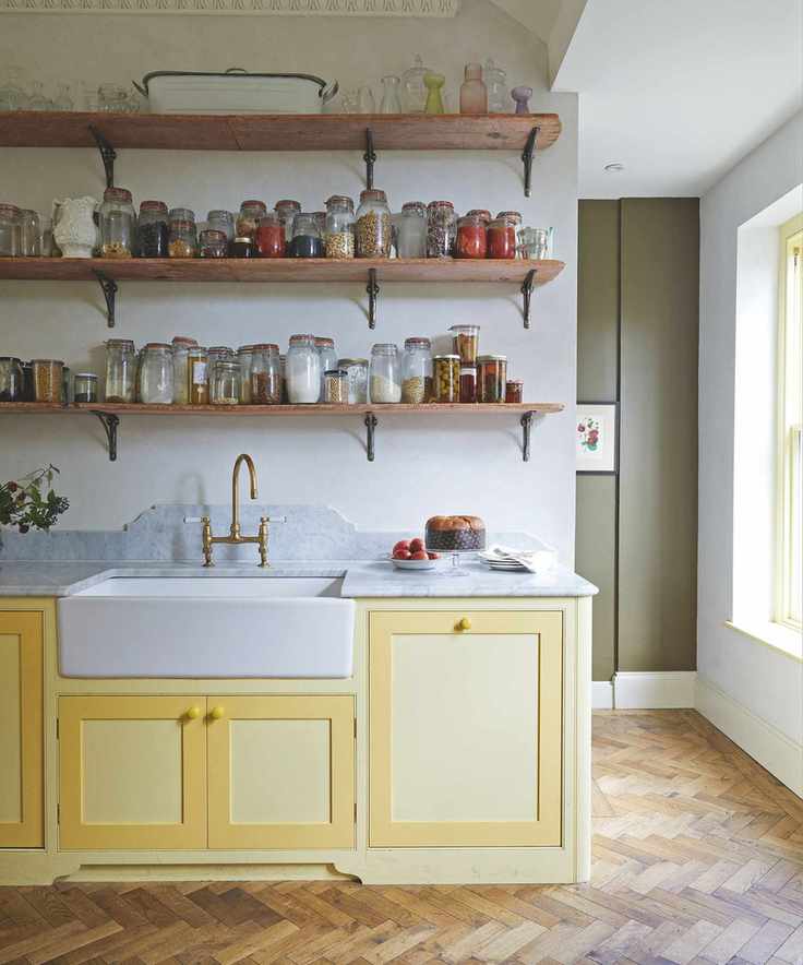 Kitchen with yellow cabinets, wooden herringbone floor and open shelves on the wall behind the sink, showing dozens of mismatched glass storage jars
