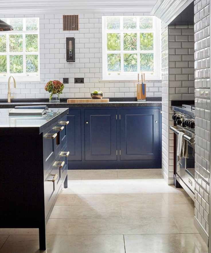 A white tiled kitchen with blue cabinets, gold hardware and faucets, and a brown shiny tiled floor