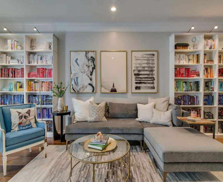 Home library with shelving, neutral L-shaped sofa, blue armchair, wood floor, glass coffee table, neutral patterned rug, neutral wall with artwork