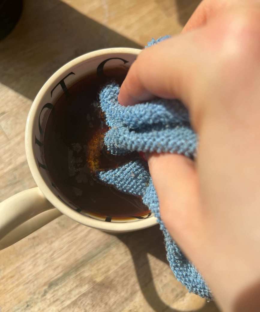 A hand dipping a blue microfiber cloth in a mug of tea