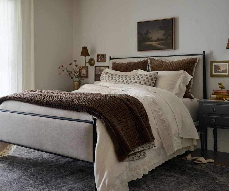 Bedroom with iron bed frame, neutral bedding with brown throw, vintage landscape on wall, side table with books and sconce