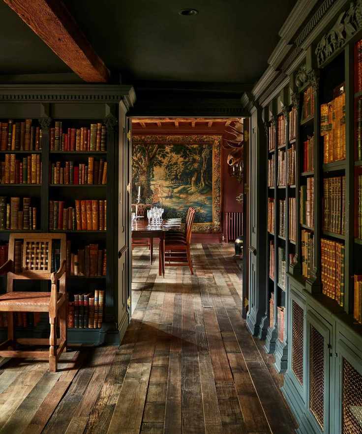 A dark-toned library with floor-to-ceiling green bookshelves filled with leather-bound books, leading into a formal dining room with a large wall tapestry and a wooden table