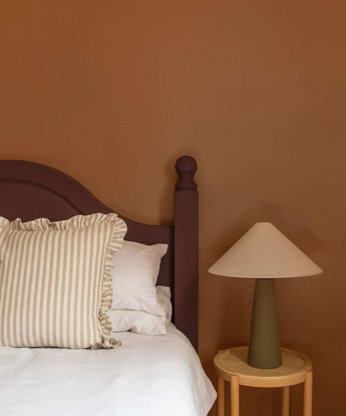 bedroom with dark orange wall, dark red bed frame and white bedding with neutral striped cushion and small table with modern lamp