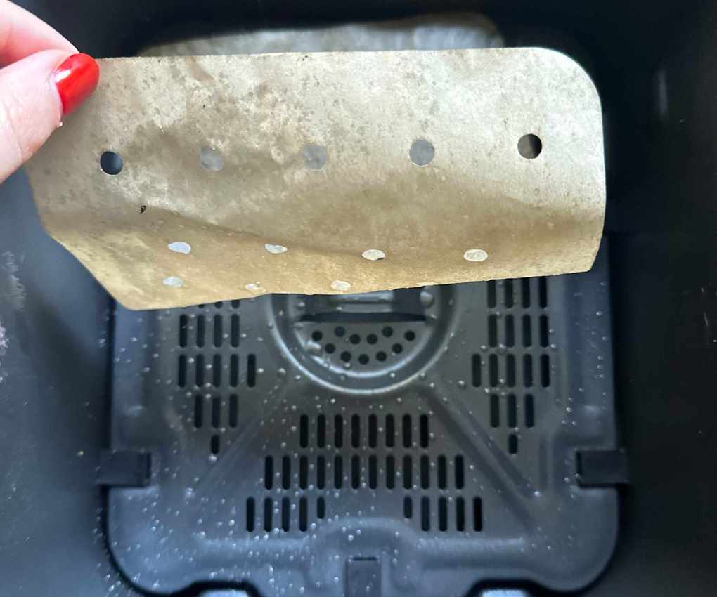 Someone with red painted nails picking up a greasy perforated paper air fryer liner from a black air fryer basket.