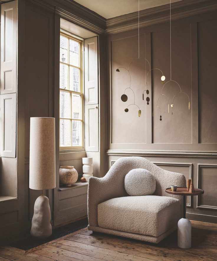 A brown panelled living room with hardwood floors, a boucle chair, large windows with shutters, a white lamp and circular table. Hanging from the ceiling is a gold mobile.