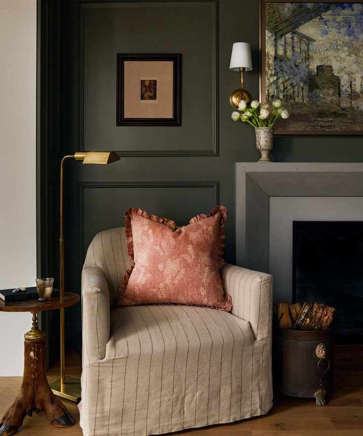 dark green living room with paneled walls, a grey fireplace with painting above, and a reading corner with a striped natural slipcovered chair with a side table and floor lamp
