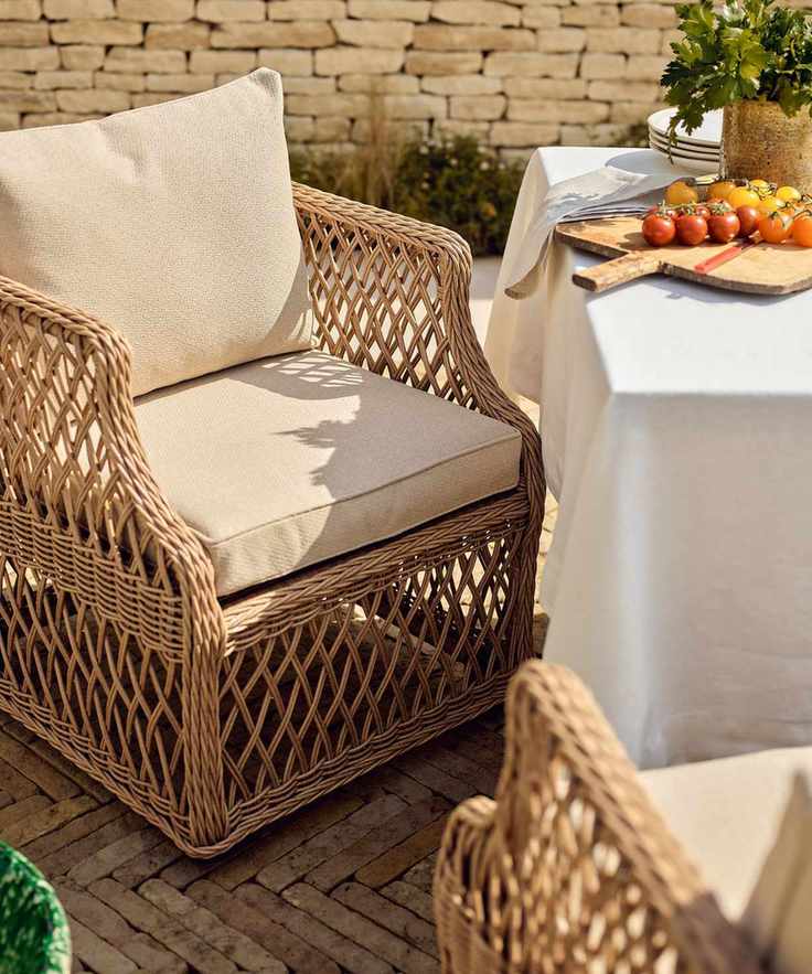 A wooden wicker outdoor armchair with cream upholstered cushions beside a garden table covered with a white cloth.